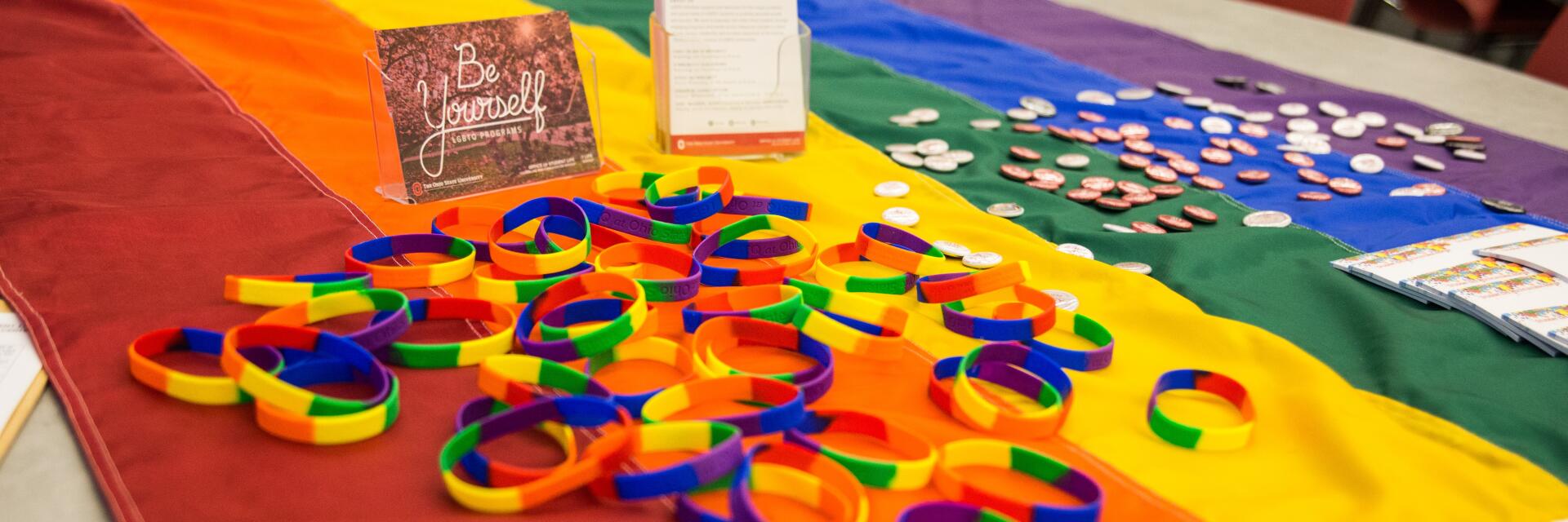 A table with a rainbow flag tablecloth, pamphlets, buttons, and rainbow rubber bracelets